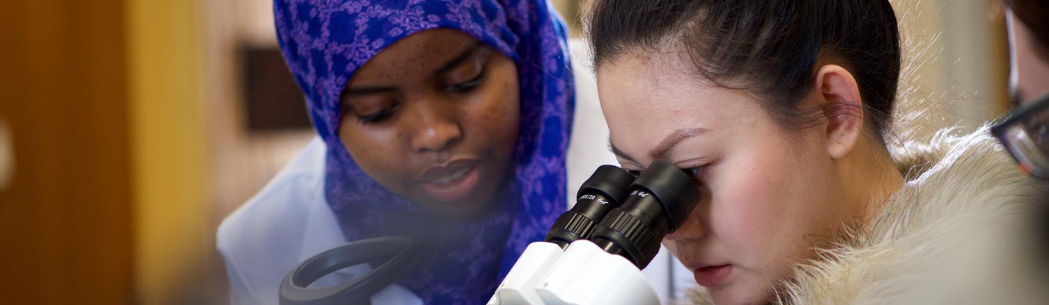 A woman looks through a microscope; another student stands next to her.