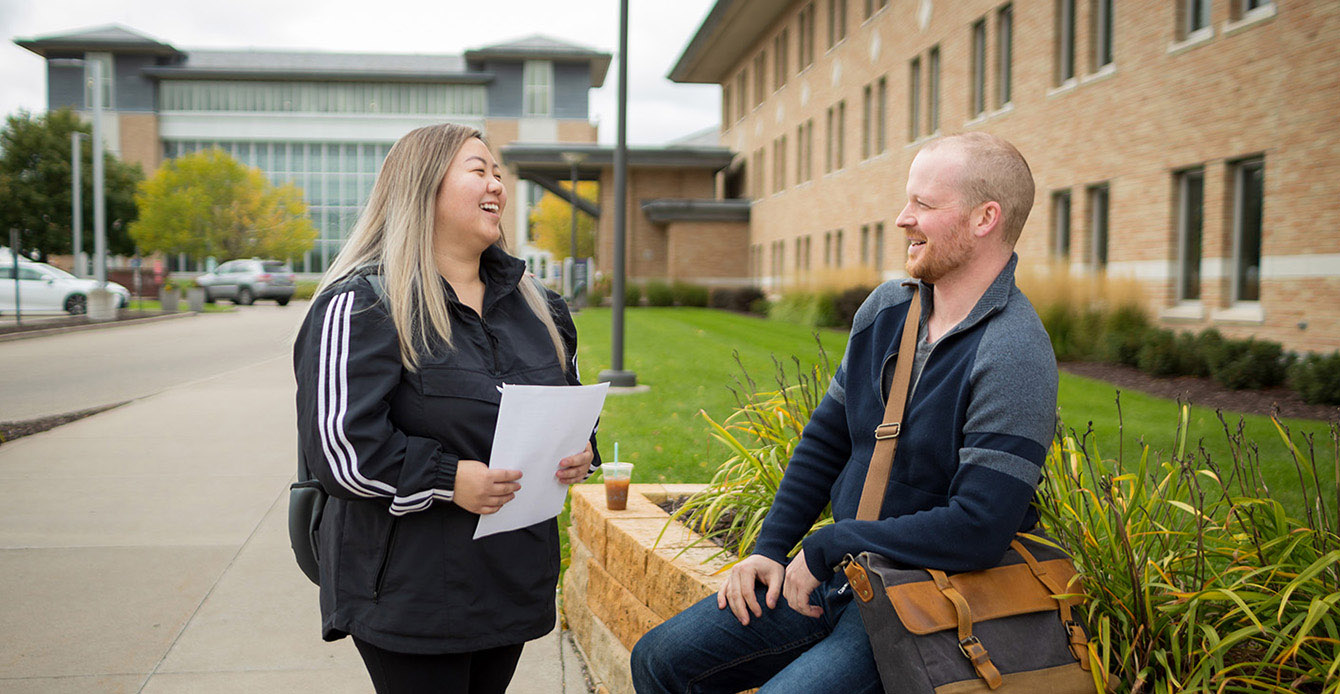 Two Metro State students talk outside on the Saint Paul campus, with campus buildings and greenery in the background.