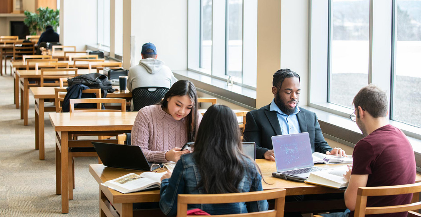 Four Metro State students study together at a table in the Library and Learning Center with textbooks and laptops open.
