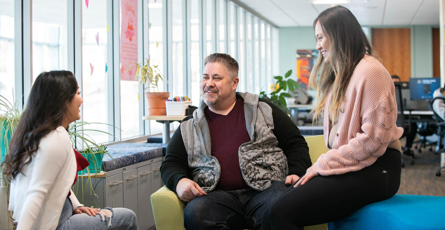 Three Metro State students talk together in a bright campus seating area near large windows in the Student Center.