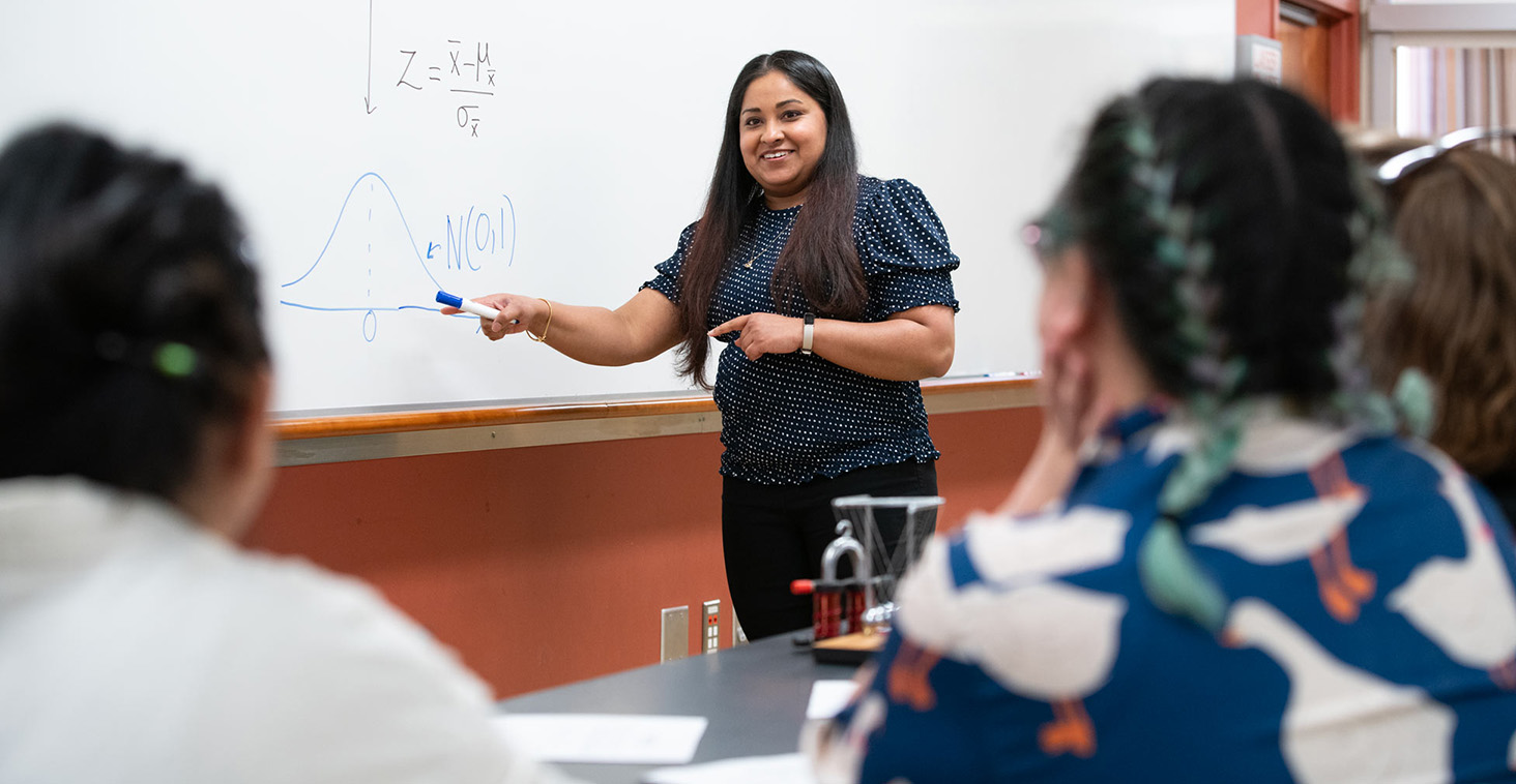 A Metro State faculty member teaches a statistics lesson at a whiteboard while students observe and take notes.