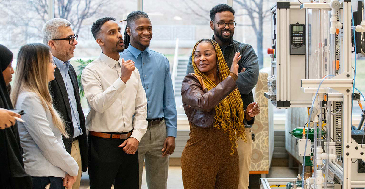 Metro State cybersecurity students and faculty gather around a water process controller, used to simulate cyberattacks against water-sector systems, in the Science Education Center atrium.