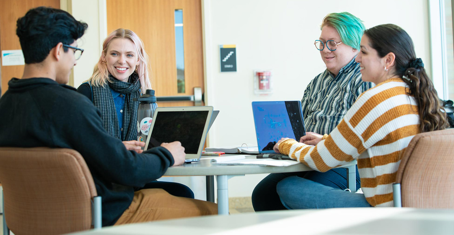 Four Metro State students talk around a table in a campus study space in the Science Education Center with laptops open.