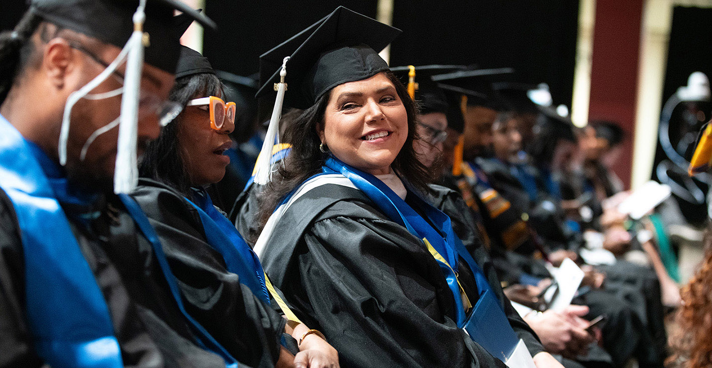 Metro State graduates in caps and gowns sit together during commencement, with one graduate smiling toward the camera.